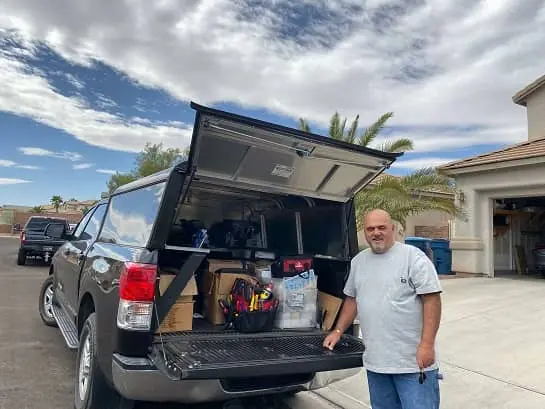 Locksmith Las Vegas man smiles by black pickup with camper shell and loaded bed in suburban driveway.