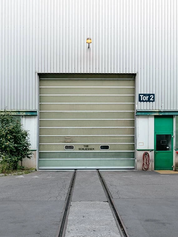 Gray industrial commercial building exterior with a closed, light beige sectional overhead door and rail tracks in the foreground.