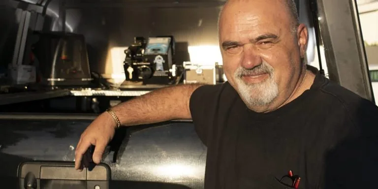 A partial view of a person with a grey beard wearing a black shirt is visible on the left side of the image, against a dark background.